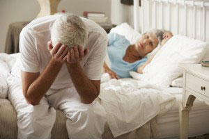 elderly man resting his head in his hands while his wife lies in bed in the background